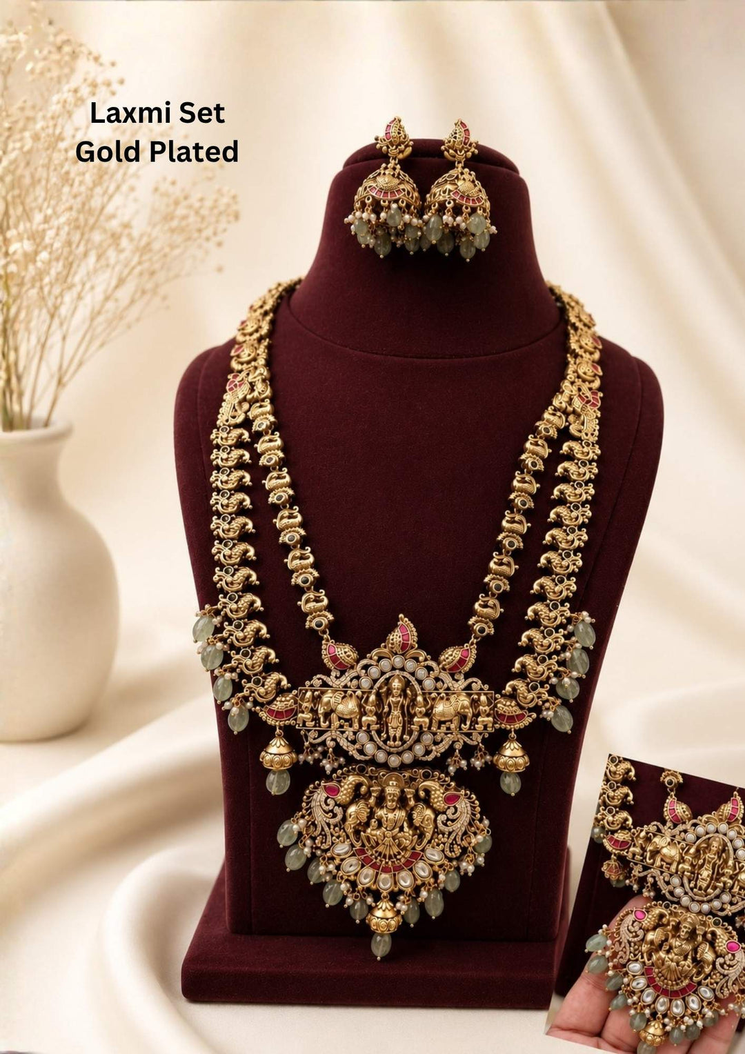 gold plated temple jewelry haram set featuring a grand double-layered long chain decorated with miniature peacock motifs. The strands lead to a structured horizontal panel depicting Goddess Lakshmi with elephants, connecting down to an intricate, heavy floral medallion pendant of a seated deity. The set is highlighted with red ruby accents and hanging light green beads. Includes matching dome jhumka earrings. Perfect for weddings and festivals with skin-friendly premium quality