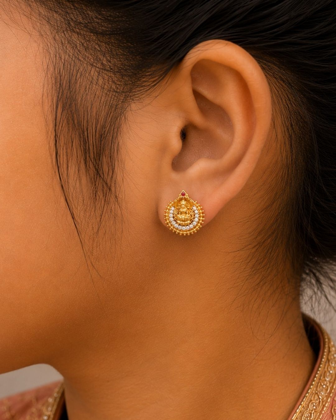 Traditional Laxmi gold plated stud earrings . The circular ear studs feature a highly detailed embossed figure of Goddess Lakshmi sitting on a lotus in the center. The idol is semi-circled by shimmering white CZ American diamond stones, a beaded gold border, and a small red ruby stone at the very top tip. This temple jewelry piece provides an elegant ethnic look suitable for puja festivals, weddings, and traditional family occasions. 