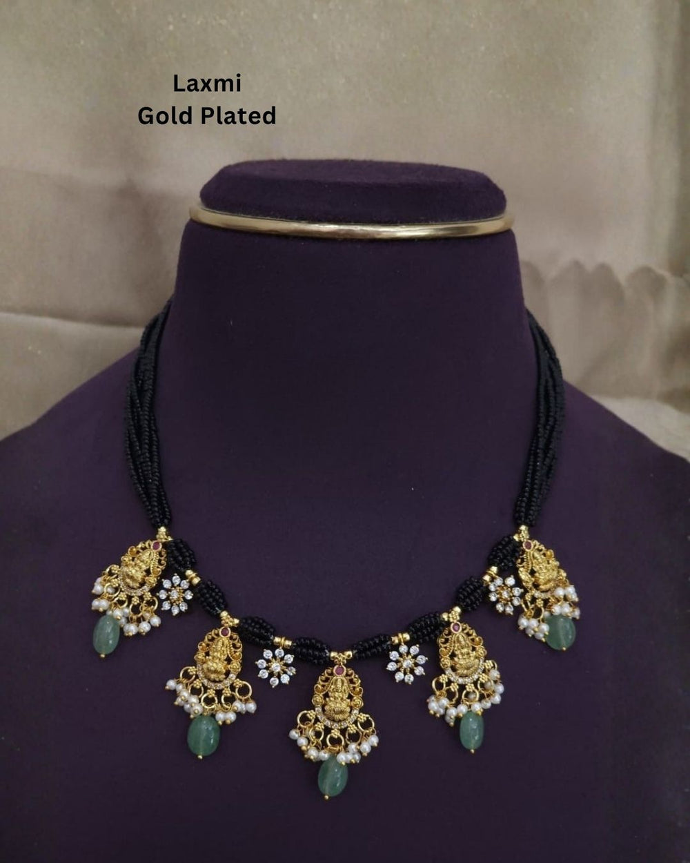 A gold-plated Laxmi necklace displayed on a purple velvet mannequin bust. The necklace features a black beaded collar with five ornate gold-plated pendant drops decorated with white faux pearls and green stone accents.