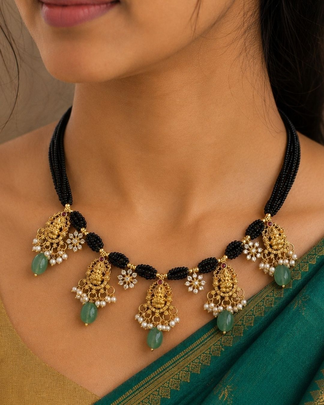 A close-up of a woman wearing a traditional Indian black bead and gold-plated necklace with green stone drops and white pearl accents, paired with a teal sari.