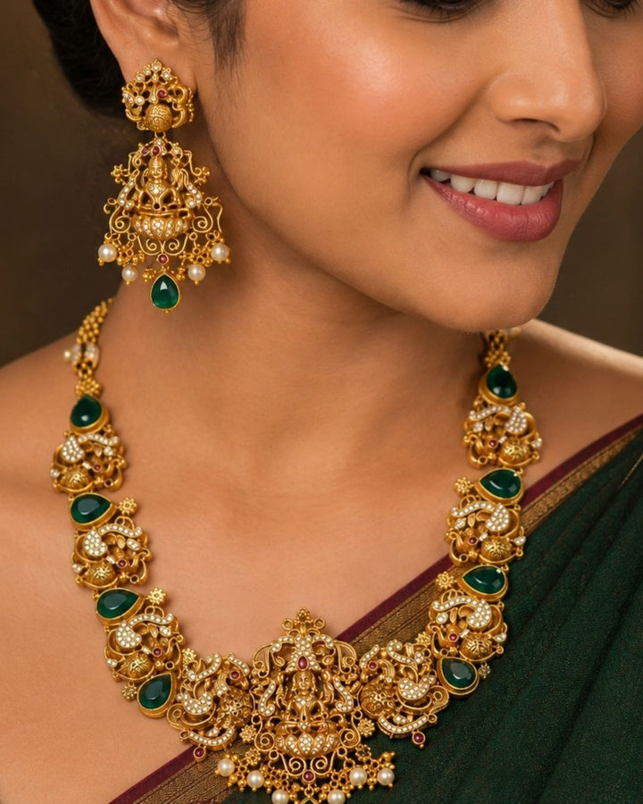 A close-up of a woman wearing an ornate gold-toned traditional necklace and matching dangle earrings with green stone accents and small pearl-like details, against a warm brown background.