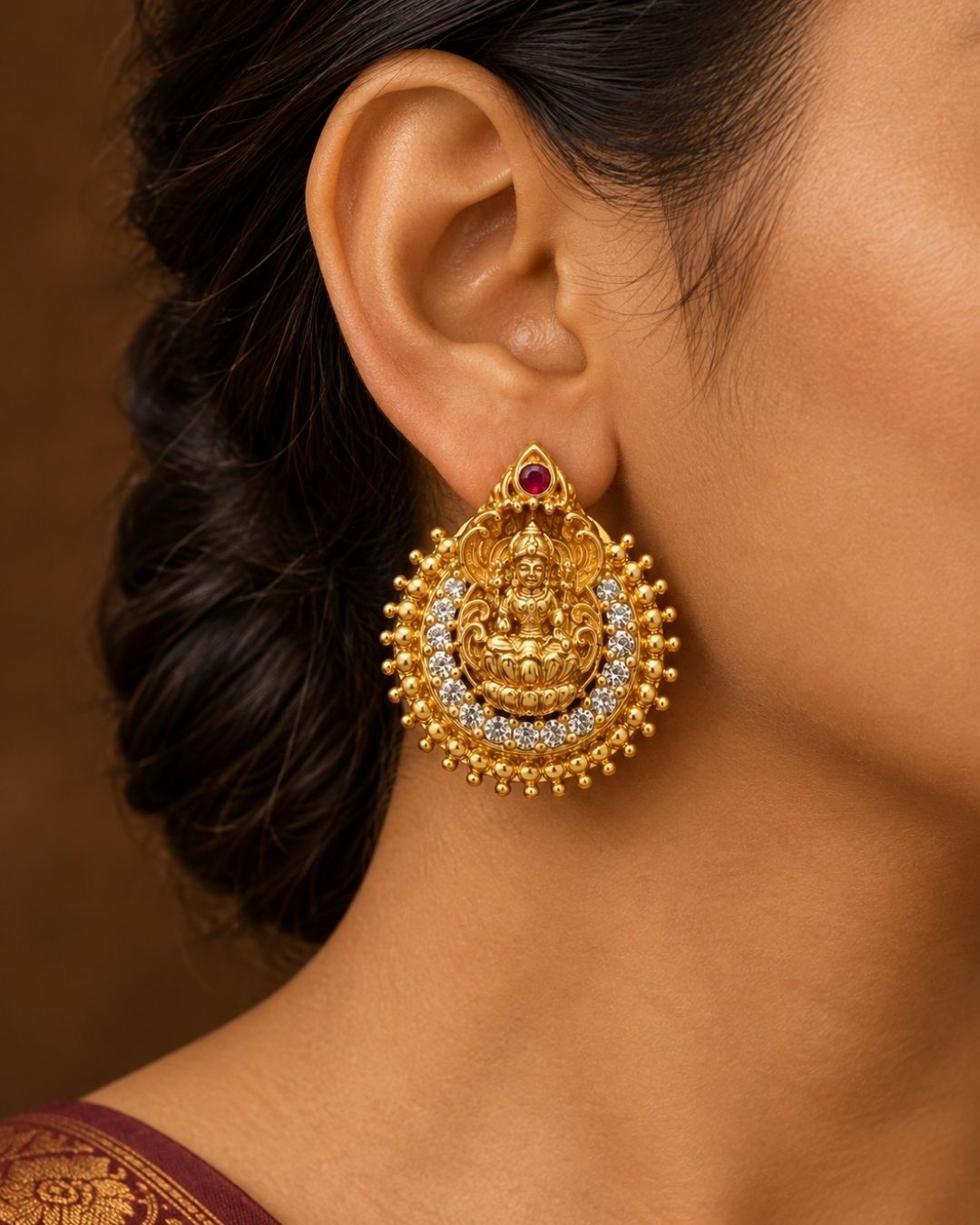 Close-up of South indian women ear wearing a Traditional Laxmi gold plated stud earrings . The circular ear studs feature a highly detailed embossed figure of Goddess Lakshmi sitting on a lotus in the center. The idol is semi-circled by shimmering white CZ American diamond stones, a beaded gold border, and a small red ruby stone at the very top tip. This temple jewelry piece provides an elegant ethnic look suitable for puja festivals, weddings, and traditional family occasions. 