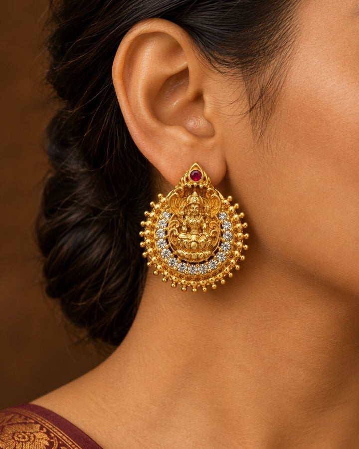Close-up of South indian women ear wearing a Traditional Laxmi gold plated stud earrings . The circular ear studs feature a highly detailed embossed figure of Goddess Lakshmi sitting on a lotus in the center. The idol is semi-circled by shimmering white CZ American diamond stones, a beaded gold border, and a small red ruby stone at the very top tip. This temple jewelry piece provides an elegant ethnic look suitable for puja festivals, weddings, and traditional family occasions. 