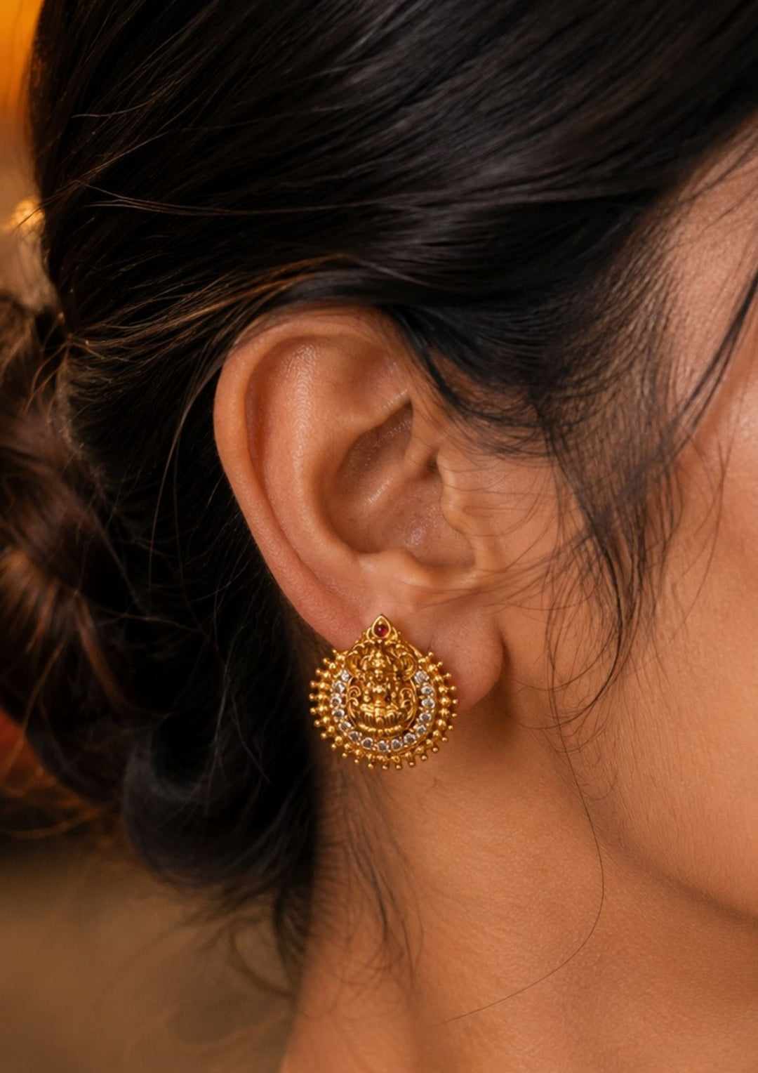 Close-up of South indian women ear wearing a Traditional Laxmi gold plated stud earrings . The circular ear studs feature a highly detailed embossed figure of Goddess Lakshmi sitting on a lotus in the center. The idol is semi-circled by shimmering white CZ American diamond stones, a beaded gold border, and a small red ruby stone at the very top tip. This temple jewelry piece provides an elegant ethnic look suitable for puja festivals, weddings, and traditional family occasions. 