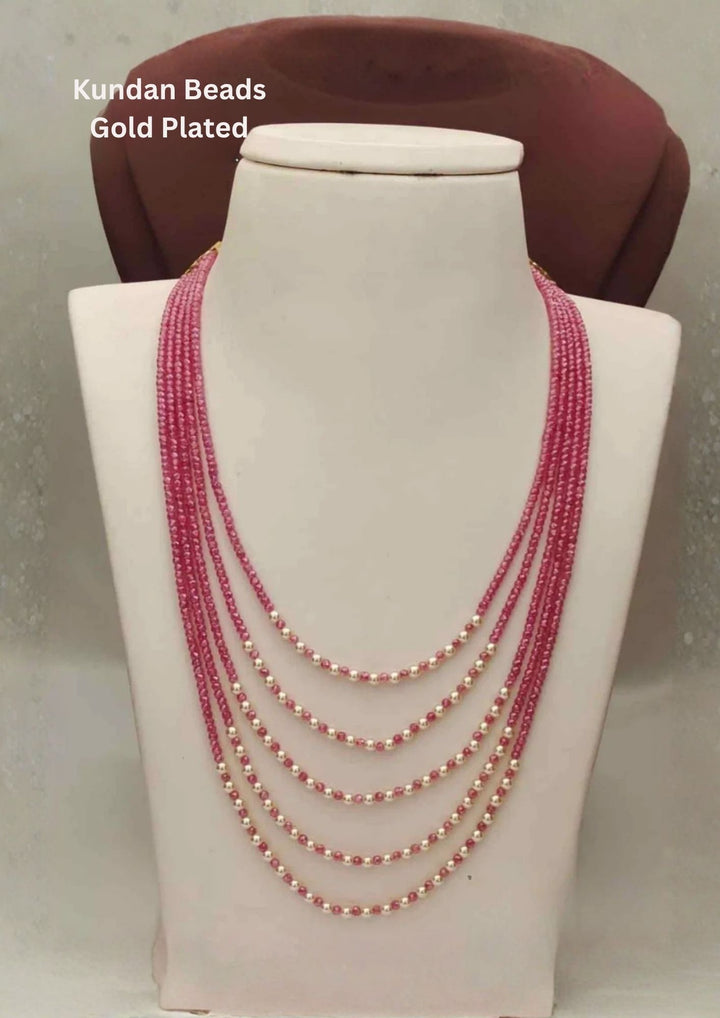 A grand five-strand layering haram necklace made of tiny, faceted ruby pink kundan seed beads. The strands are arranged in a stepped, graduated pattern on a mannequin, with the lower section of each string decorated with alternating cream-white pearls for a dual-tone contrast.