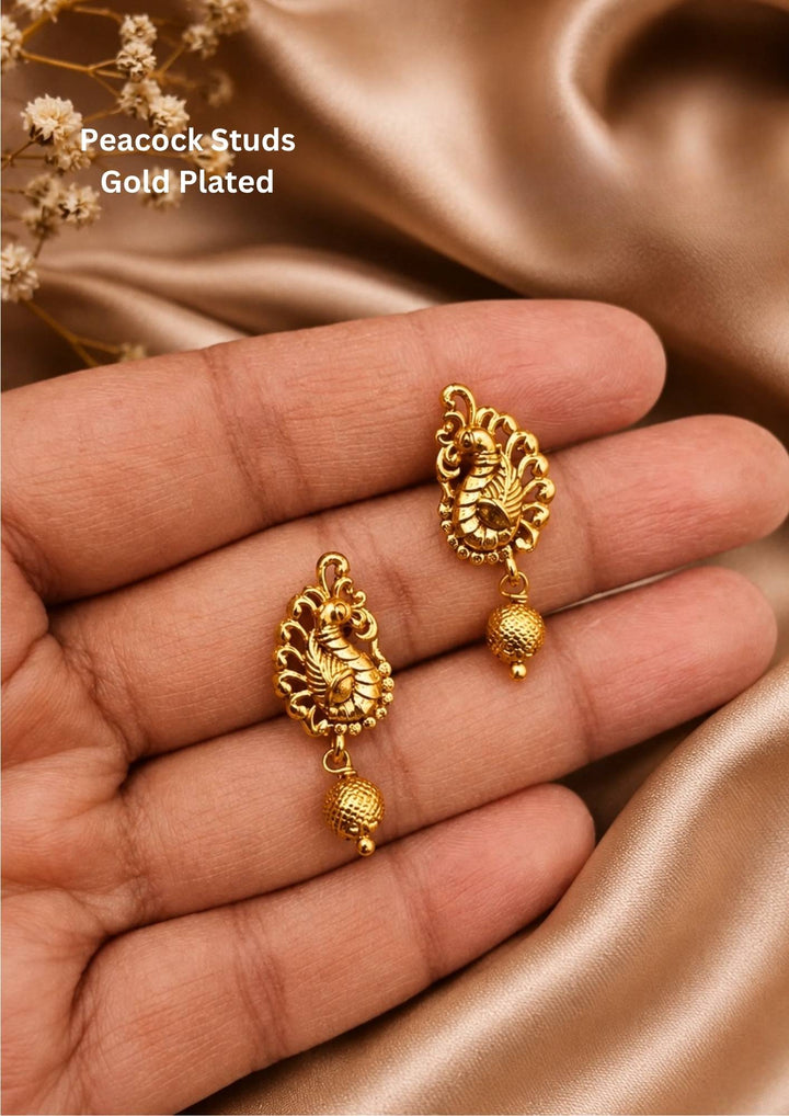Gold plated stud earrings featuring a traditional upright peacock design. The body of the peacock is crafted with clean, textured gold details, and a small, round textured gold ball hangs gently from the bottom of the bird shape, providing a simple, classic look suitable for daily wear, festivals, and family functions.