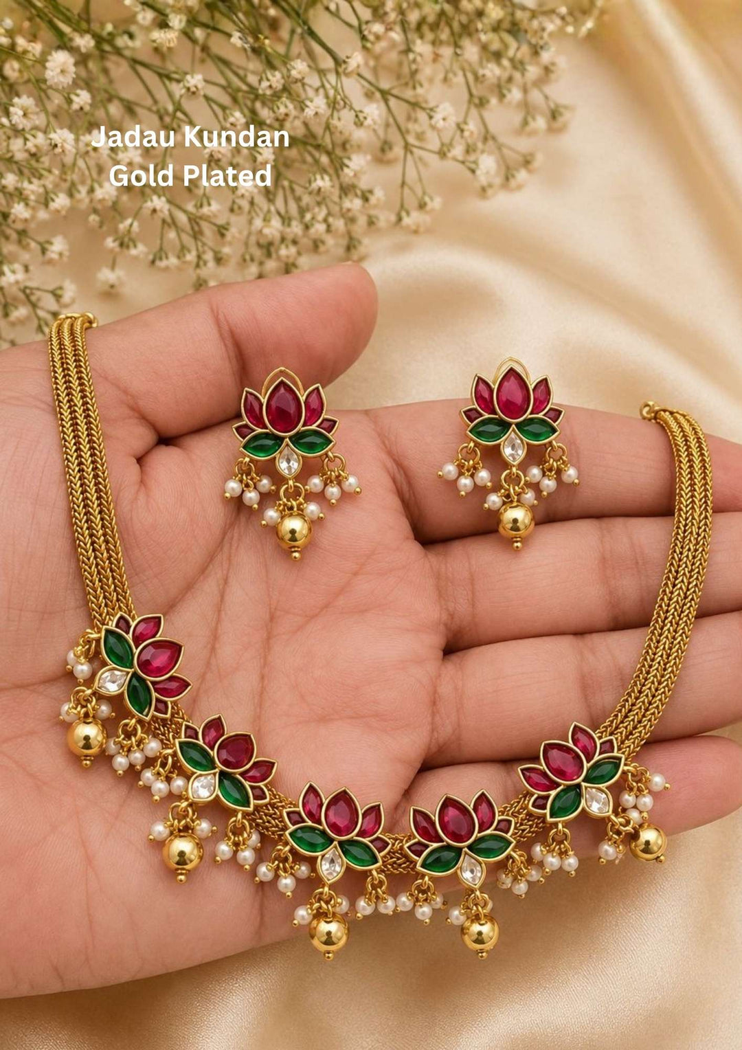 Jadau kundan gold plated lotus motif necklace set featuring a row of beautifully detailed pink ruby and green emerald gemstone lotus flowers held together by a three-strand woven gold chain. Each lotus flower station is accented with a clear kundan base stone, tiny white pearl clusters, and hanging smooth gold bead droplets, paired with matching lotus flower drop earrings that provide a rich and classy appearance suitable for weddings, festivals, receptions with skin friendly quality, anti fade polish