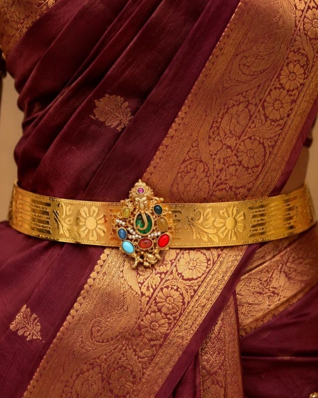 Beautiful gold plated adult hip belt featuring an intricately engraved wide band with floral designs and a central Ganesha motif surrounded by colorful green, red, blue, and white stones. Perfect for traditional wear, weddings, festive occasions, bridal styling, parties, and gifting.