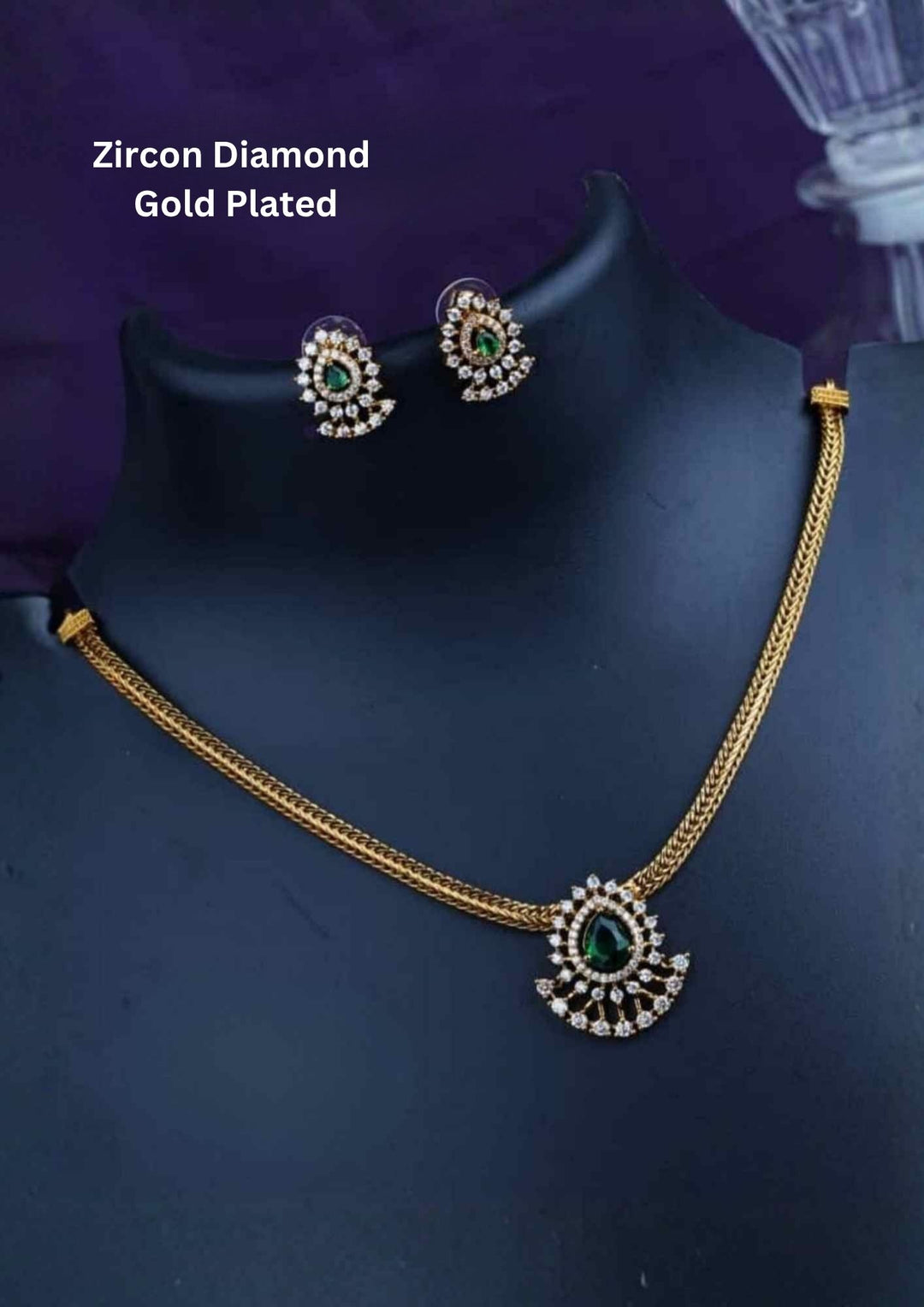 A minimalist 18k gold plated herringbone chain necklace featuring a central teardrop-shaped ruby green stone. The stone is enclosed in a halo of white pure Zircon diamonds that flares out at the bottom into a shimmering, fan-shaped scalloped fringe, paired with matching crown-set ear studs.