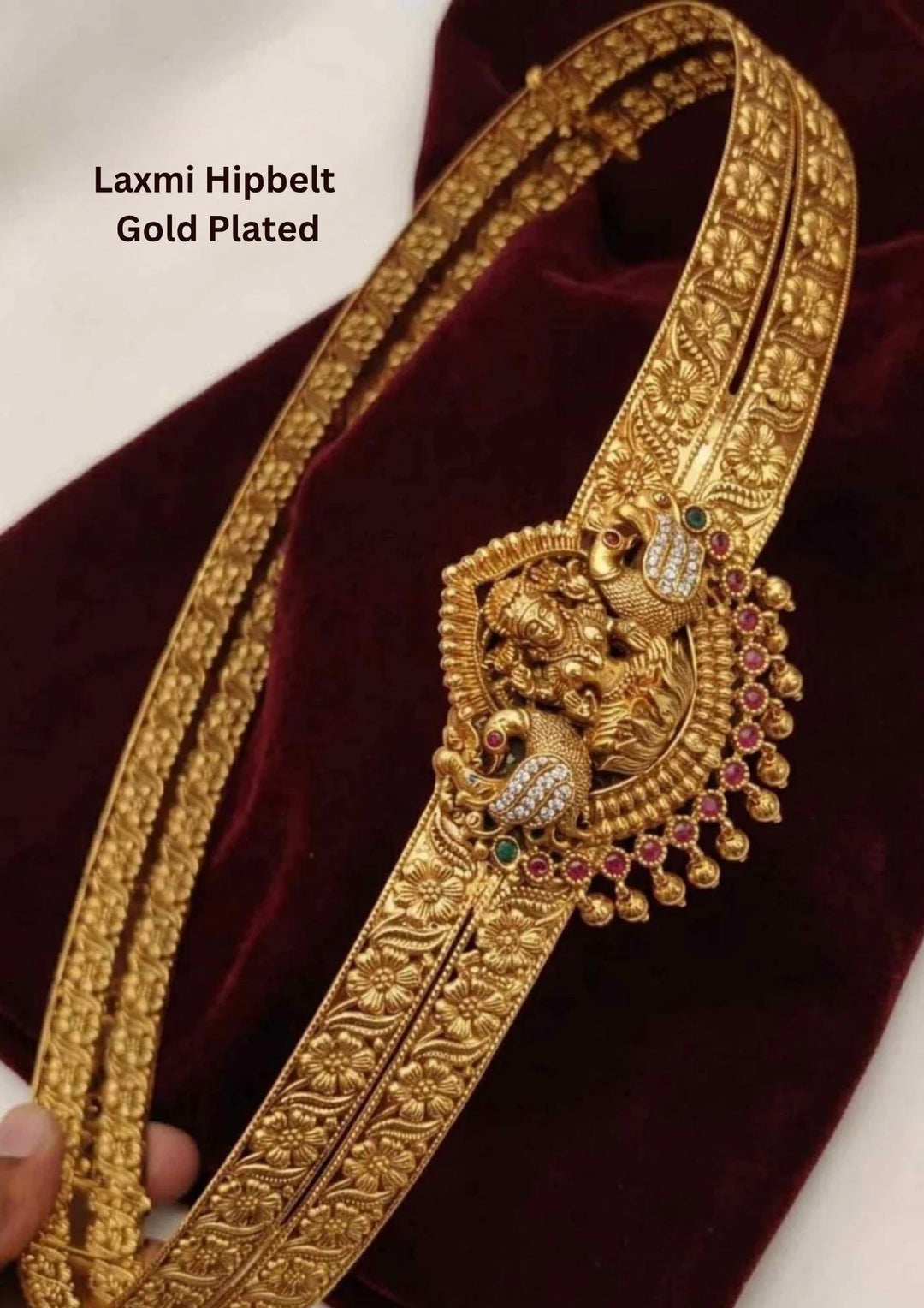 gold plated adult hipbelt vaddanam featuring two parallel flexible bands deeply embossed with repeating floral motifs. The central medallion showcases a detailed carving of Goddess Lakshmi flanked by twin peacocks inlaid with white crystals, emerald accents, and a fringe of hanging golden beads.