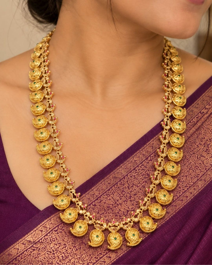 Elegant gold plated long haram featuring intricately crafted paisley motifs adorned with green and ruby stone accents, paired with matching earrings. Inspired by traditional South Indian temple jewellery, this timeless necklace set is perfect for weddings, bridal wear, festivals, and cultural celebrations.