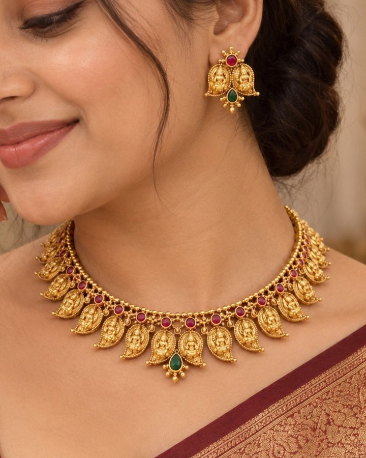 Elegant gold plated Lakshmi Devi necklace set featuring multiple detailed temple motifs, red and green stone embellishments, a central green teardrop pendant, and matching earrings, perfect for festive and traditional occasions.