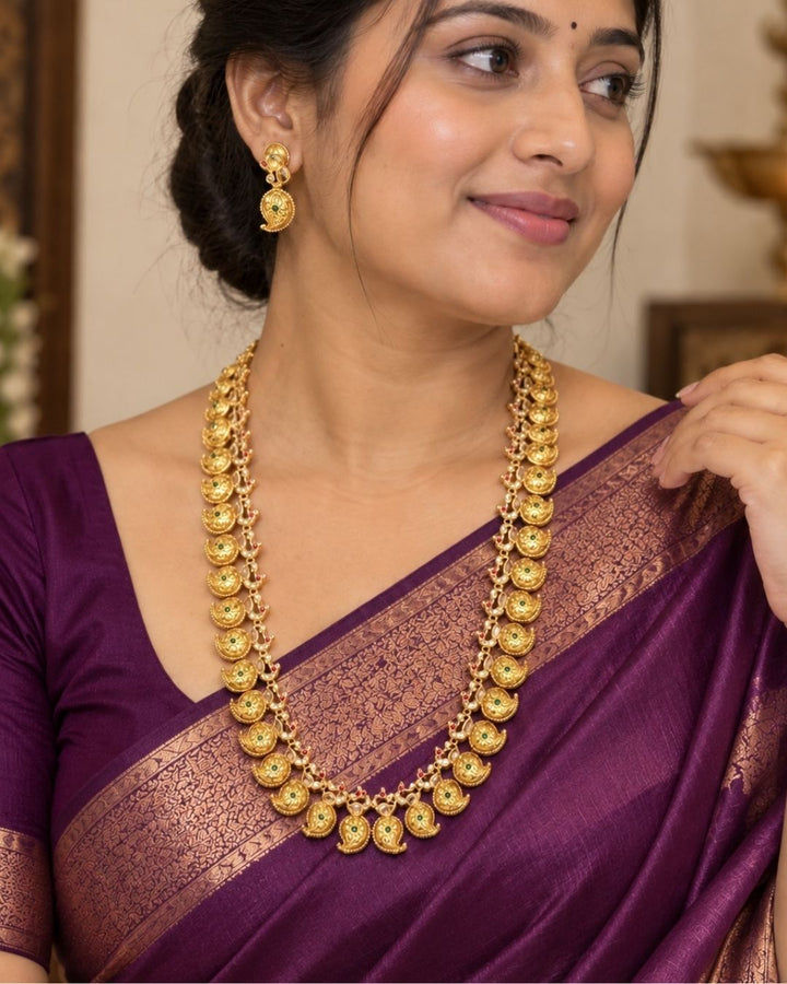 Elegant gold plated long haram featuring intricately crafted paisley motifs adorned with green and ruby stone accents, paired with matching earrings. Inspired by traditional South Indian temple jewellery, this timeless necklace set is perfect for weddings, bridal wear, festivals, and cultural celebrations.