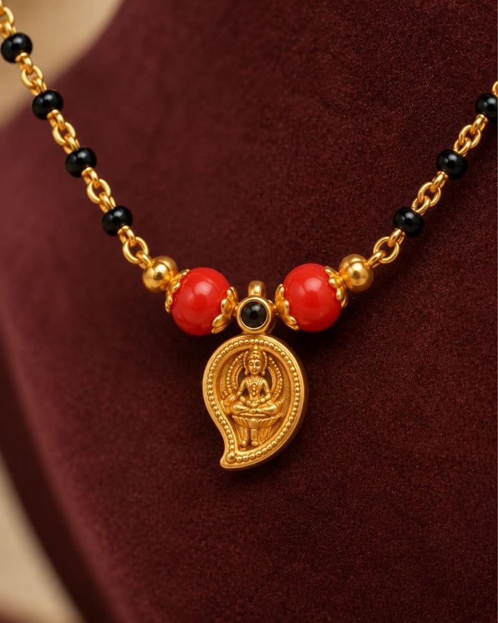 A gold-plated Mangalsutra necklace with red beads and a small gold pendant, displayed on a dark burgundy jewelry bust. Text in the upper left reads &