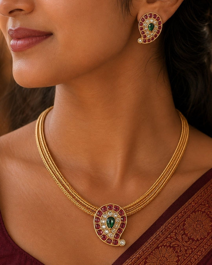 Beautiful Jadau Kundan gold plated pendant necklace set featuring a paisley motif with ruby red stones, a green center stone, sparkling kundan work, and matching earrings. Perfect for weddings, festivals, temple visits, traditional outfits, family functions, and premium gifting.