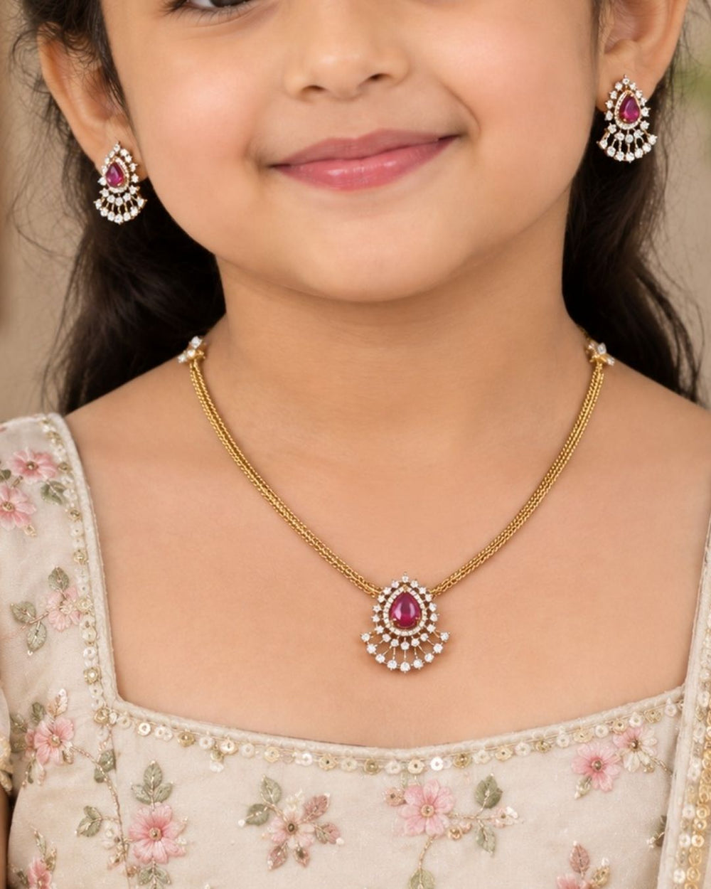 A minimalist 18k gold plated herringbone chain necklace featuring a central teardrop-shaped ruby pink stone. The stone is enclosed in a halo of white pure Zircon diamonds that flares out at the bottom into a shimmering, fan-shaped scalloped fringe, paired with matching crown-set ear studs.