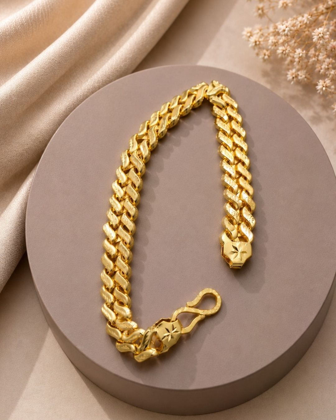 A chunky gold Cuban link chain bracelet with a star-patterned clasp, displayed on a round taupe pedestal with beige fabric and dried flowers in the background.