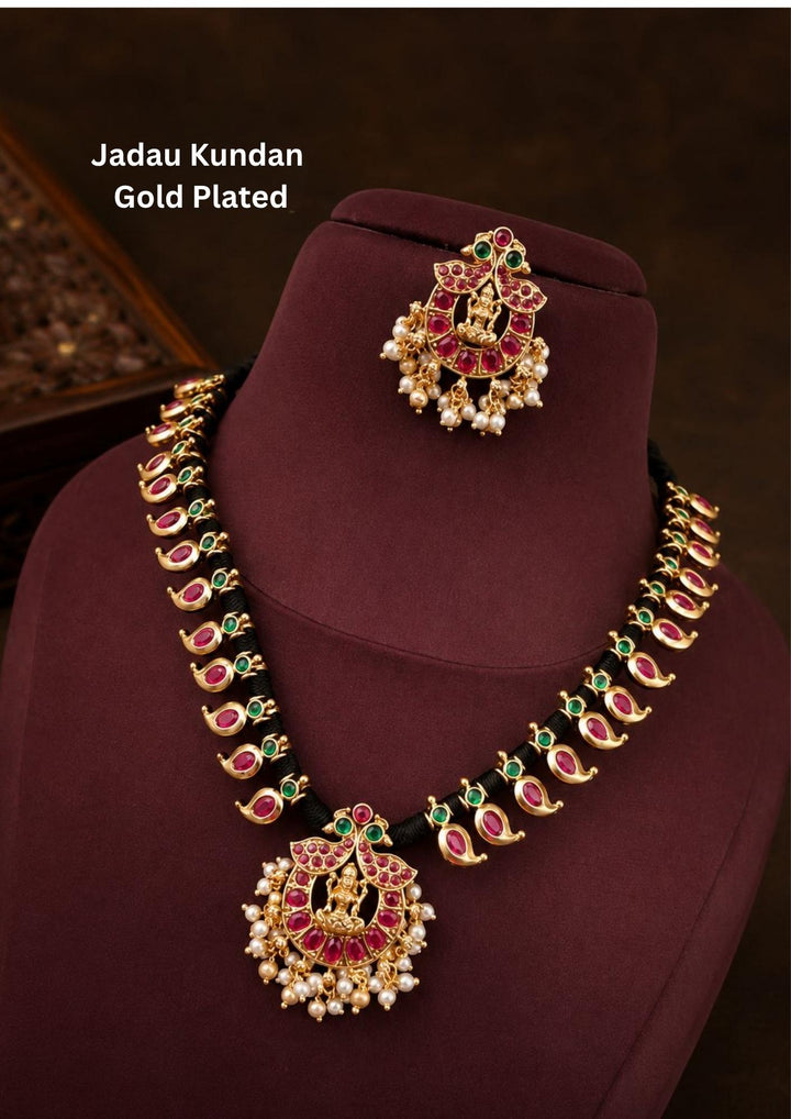 Gold plated Jadau Kundan short necklace and matching maang tikka set featuring a black thread cord lined with continuous gold paisley mango motifs inset with pink ruby stones and small green emerald spacers. The central crescent-shaped chandbali pendant and matching tikka are adorned with pink stones, clear kundan crystals, an embossed center figurine of Goddess Laxmi, suitable for fesfor festive wear and traditional occasions.