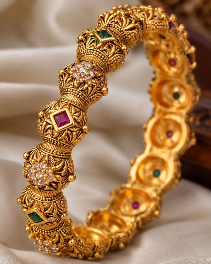 Close up of a south indian women hand wearing Traditional gold plated temple bangles showcasing ornate antique detailing, ruby pink square stones, emerald green accents, and floral zircon motifs. Perfect for bridal wear, festive occasions, and ethnic celebrations, offering a luxurious heritage-inspired look.