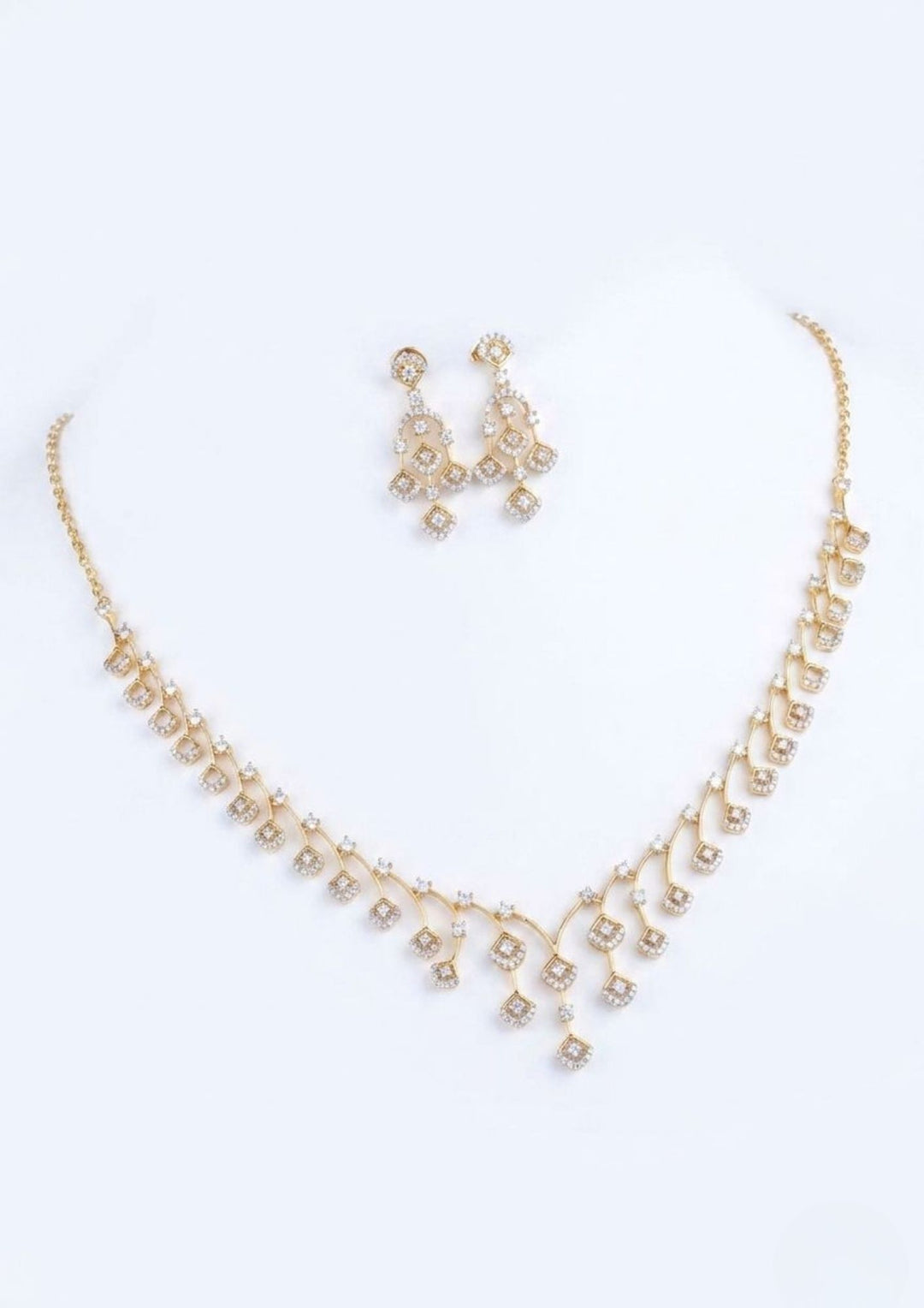 A beautiful and thin gold-plated necklace with matching earrings on a clean white background. The necklace has a V-shape design made of small, shiny white diamond-like stones. It has tiny, hanging square shapes at the bottom that look very elegant. The matching earrings have the exact same small square design and hang gently.