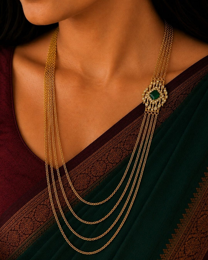 A close-up of a woman wearing a traditional Indian outfit, featuring a deep maroon saree with gold zari embroidery and a long, multi-strand gold necklace with red stone accents, along with a matching gold earring.
