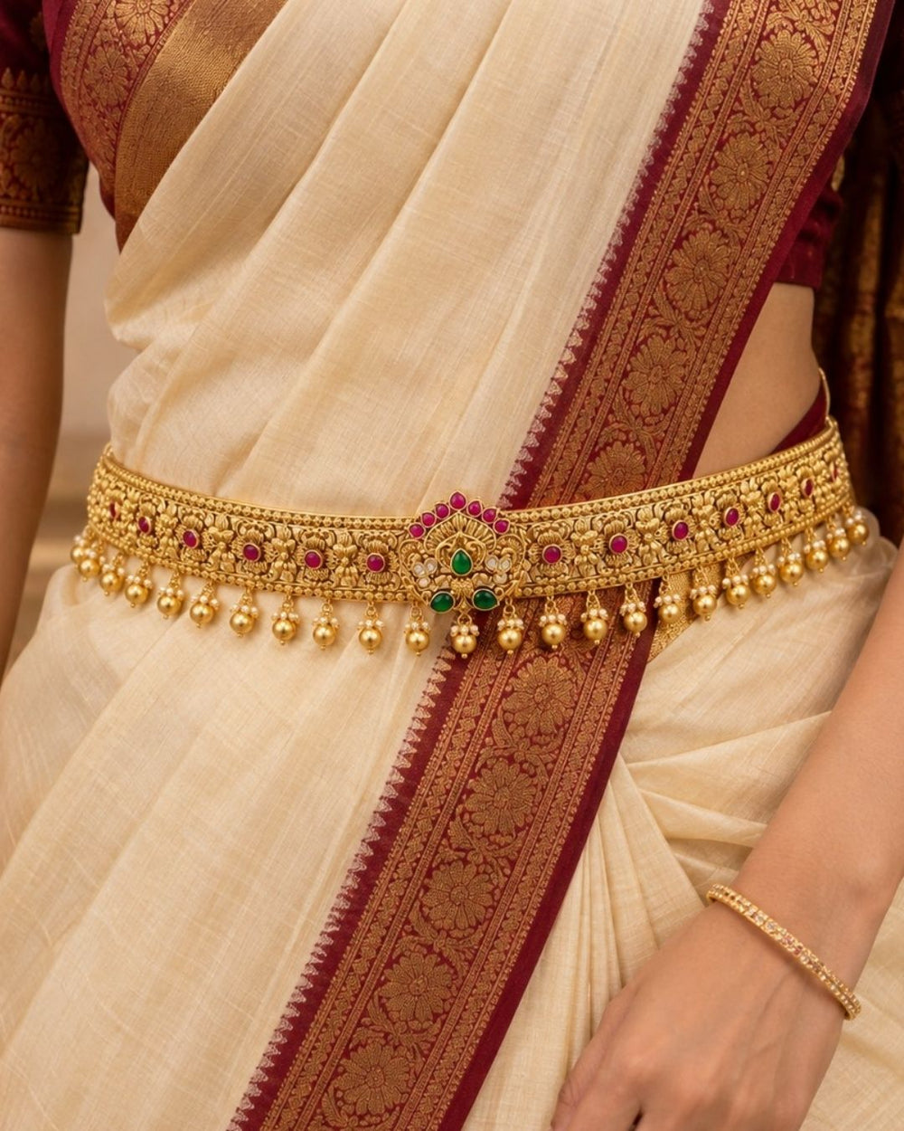 Premium gold plated designer hip belt featuring detailed floral motifs, ruby-colored stone accents, green stones at the center, and elegant hanging gold beads. Perfect for weddings, festive occasions, traditional wear, bridal styling, parties, and gifting.