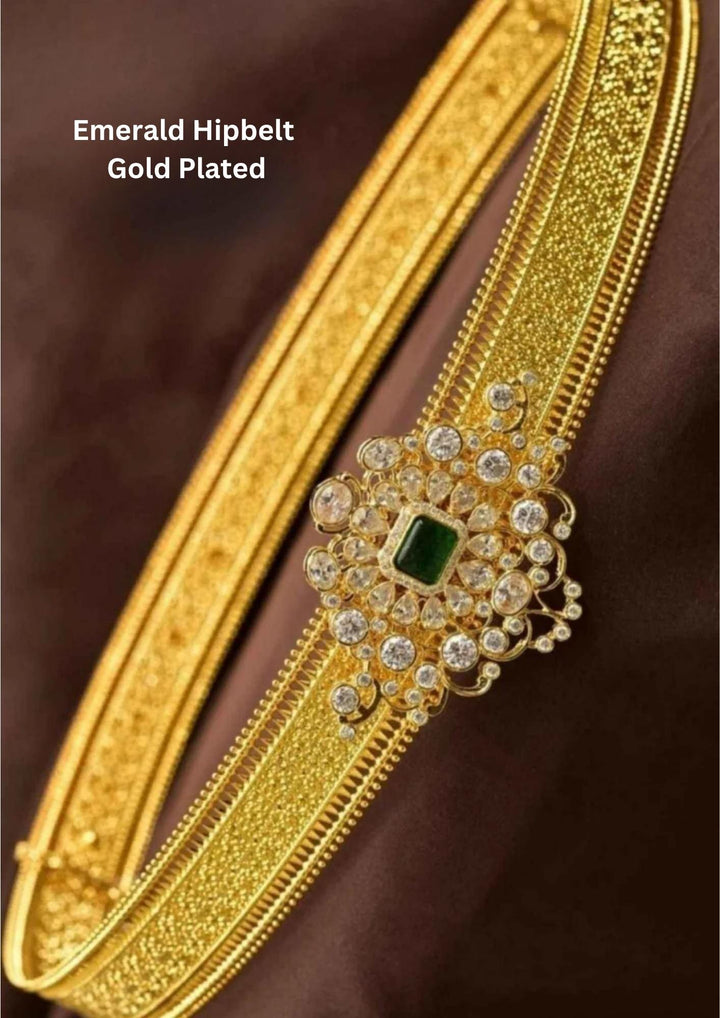 A gold plated traditional adult hipbelt (vaddanam) featuring a wide, flexible band with a shimmering stardust textured finish. The central statement ornament is a large, ornate filigree frame heavily encrusted with white zircon stones, anchoring a rich rectangular emerald green core gemstone.