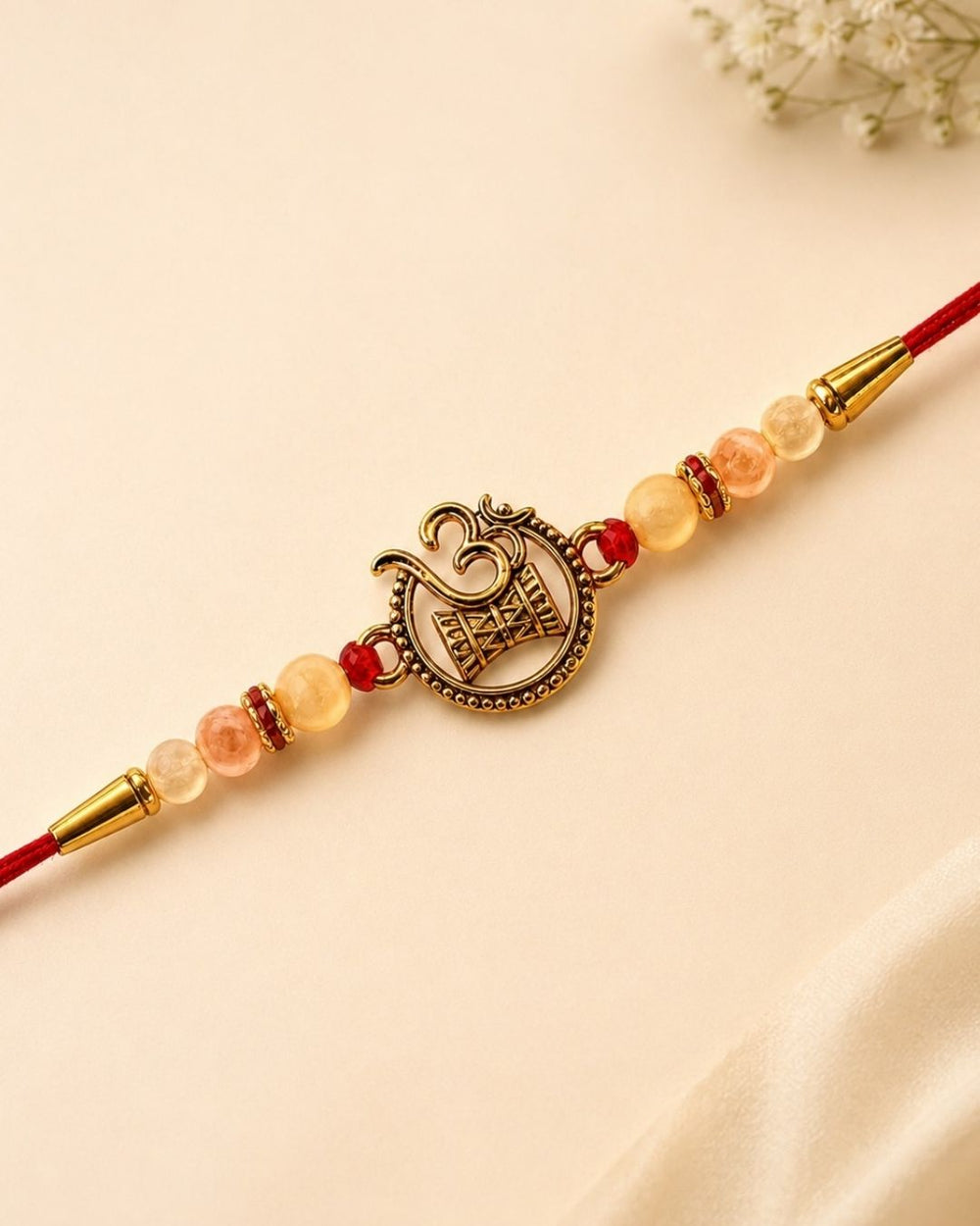 A red cord Rakhi bracelet with a central bronze Om symbol charm, accented by peach and red beads, gold spacer rings, and gold end caps, displayed on a light beige satin background.