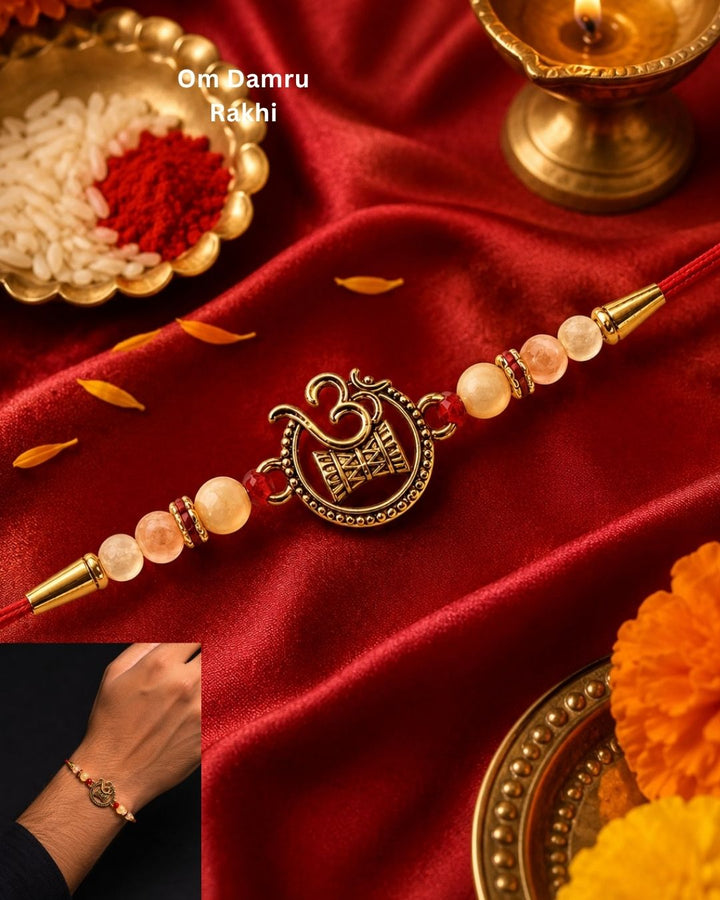 A red Rakhi bracelet with a gold-toned Om Damru charm and pinkish-beige beads, displayed on red satin with decorative brass dishes and marigold flowers. A small inset shows the bracelet worn on a person&