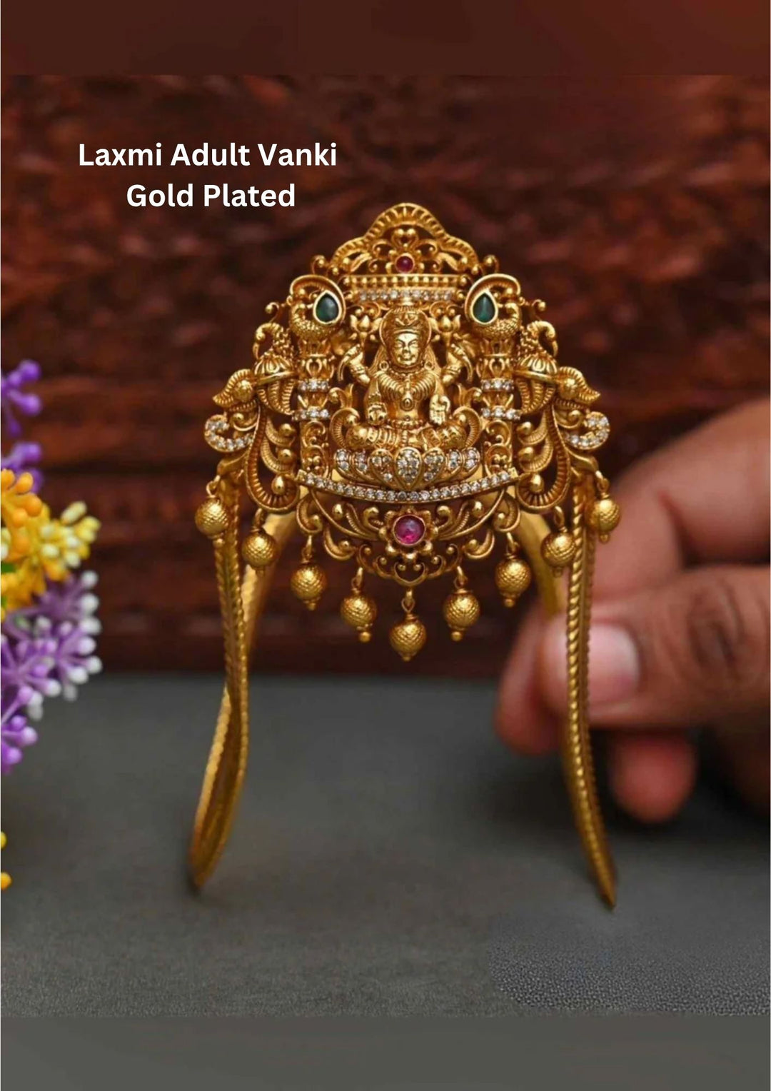 An ornate, traditional gold plated adult vanki (armlet) designed with an inverted V-shape frame. The central shield features a detailed carving of Goddess Lakshmi on a lotus, paved with white zircon crystals, flanked by twin peacocks with green emerald eyes, and finished with hanging golden beads.