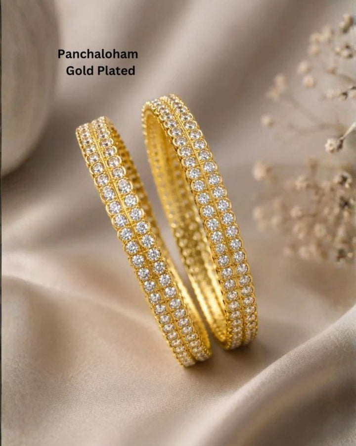 A pair of premium gold-plated bangles meticulously studded with two parallel rows of brilliant, round-cut clear zircon stones. Each crystal is secured in a classic prong setting along the outer rim of the band, creating a continuous, shimmering diamond-like effect against the rich, polished gold finish suitable for weddings, poojas and bridal occasions.