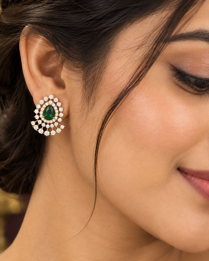 A minimalist 18k gold plated herringbone chain necklace featuring a central teardrop-shaped ruby green stone. The stone is enclosed in a halo of white pure Zircon diamonds that flares out at the bottom into a shimmering, fan-shaped scalloped fringe, paired with matching crown-set ear studs.
