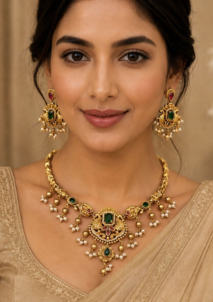 Woman wearing a An exquisite Jadau Kundan gold plated necklace features a rigid gold-engraved band transitions into twin peacock motifs framing a central rectangular green emerald stone, accented by delicate hanging pearl clusters and matching drop earrings.