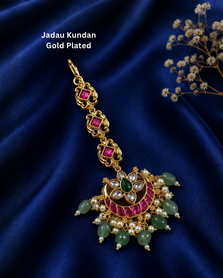 Beautiful traditional gold plated maang tikka featuring a pink stone chain, a floral-inspired circular pendant with green and pink stones, sparkling white accents, clusters of pearls, and multiple green bead drops. Perfect for weddings, festive occasions, traditional outfits, bridal styling, and special celebrations.