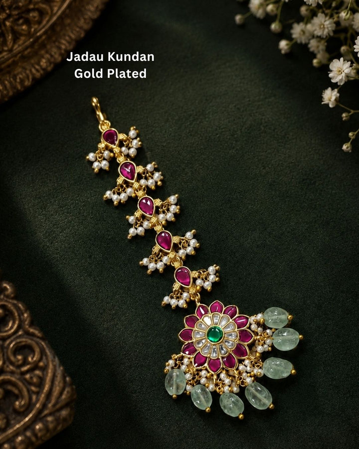 Beautiful traditional gold plated maang tikka featuring multiple pink teardrop stones along the chain, a floral pendant with vibrant pink petals and a green center stone, sparkling white stones, delicate pearl clusters, and multiple green bead drops. Perfect for weddings, festive occasions, traditional outfits, bridal styling, and special celebrations.