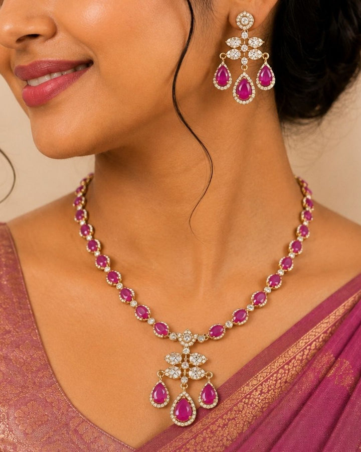 Gold-plated Indian style jewelry set featuring a necklace and matching earrings with pink teardrop-shaped stones and clear pavé accents, modeled on a woman wearing a purple saree.