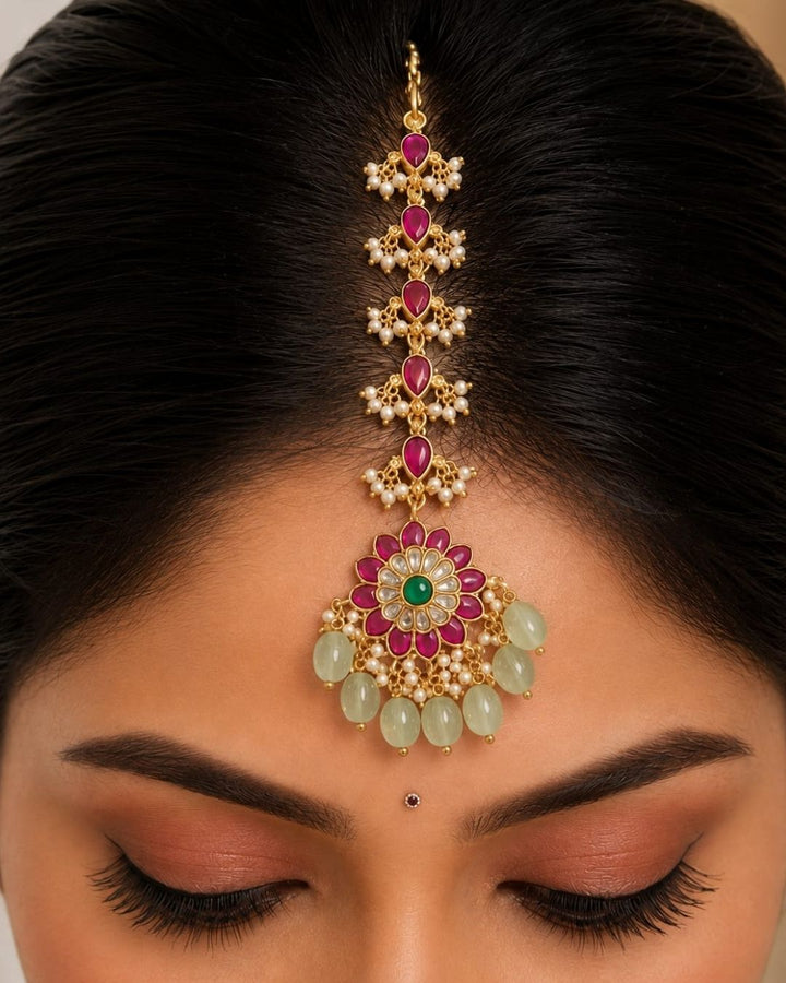 Beautiful traditional gold plated maang tikka featuring multiple pink teardrop stones along the chain, a floral pendant with vibrant pink petals and a green center stone, sparkling white stones, delicate pearl clusters, and multiple green bead drops. Perfect for weddings, festive occasions, traditional outfits, bridal styling, and special celebrations.