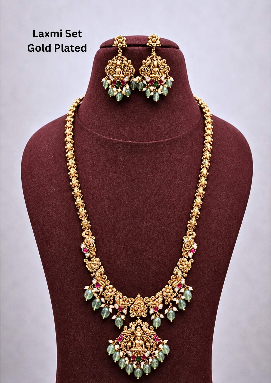 gold plated brass base temple jewelry haram set featuring a long intricate chain that transitions into floral and peacock links, centering a grand medallion pendant embossed with a seated Goddess Lakshmi motif. The piece is highlighted with pink and green stones and finished with dangling light green tumbles and pearl clusters. Includes matching grand hanging earrings. Perfect for weddings and festivals with skin-friendly premium quality