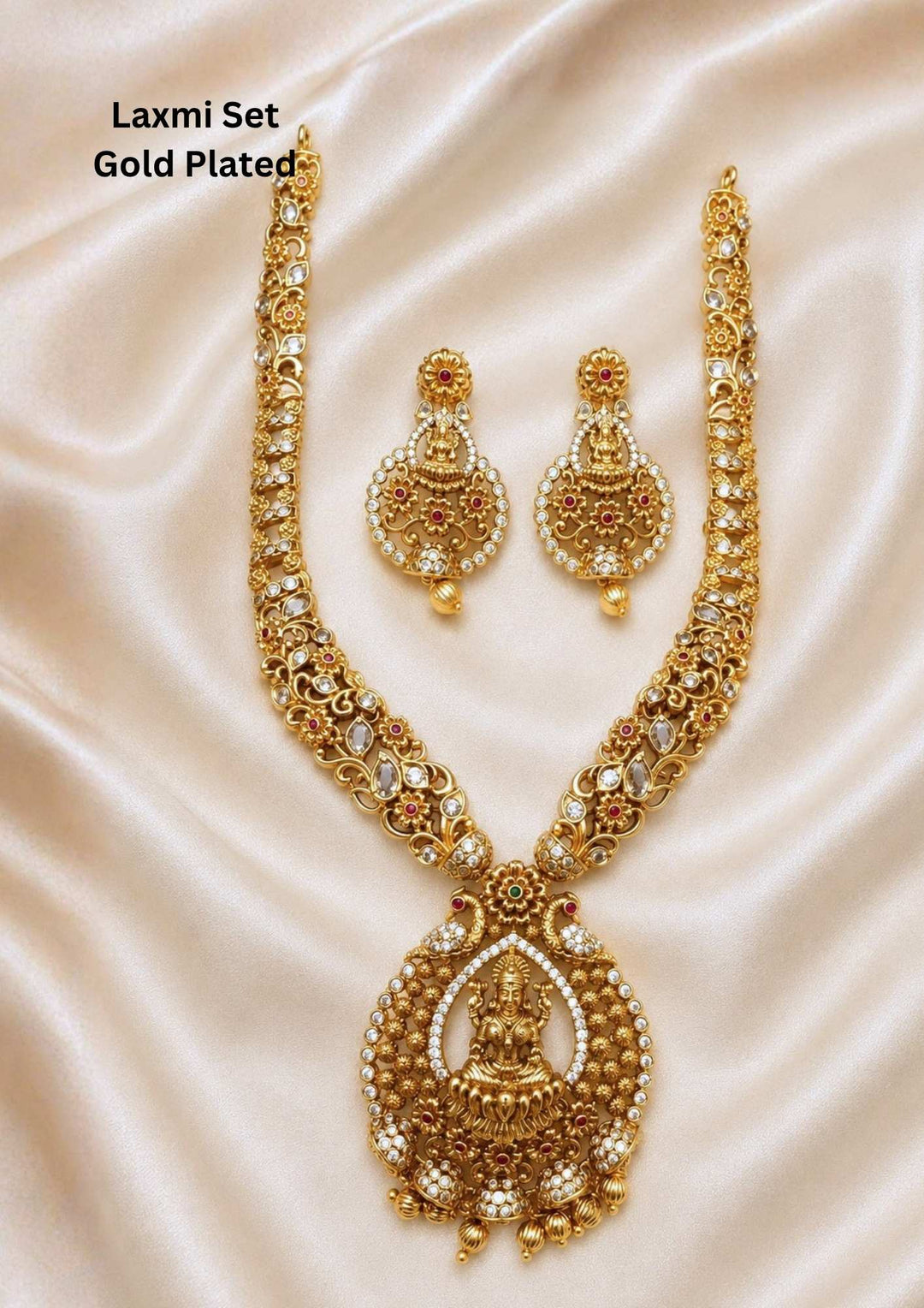 Antique gold plated temple jewelry haram set featuring a long intricate floral chain studded with small white and pink stones, leading down to a grand circular medallion pendant with a central Goddess Lakshmi motif seated on a lotus. The matching hanging earrings feature a coordinating seated Lakshmi design with gold bead drops. Perfect for weddings, festivals, and temple visits with skin-friendly premium quality, comfortable fitting.