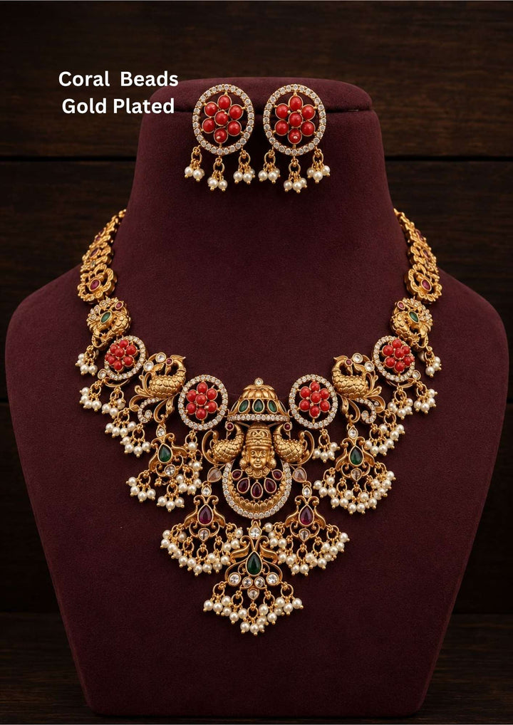 Coral beads gold plated Goddess Lakshmi long haar featuring a grand antique gold temple pendant with a central seated Lakshmi motif flanked by dual peacocks, red rubies, and green emeralds, suspended from a heavy four-strand chain of orange coral beads and white pearls. This long haram includes matching layered coral bell jhumka earrings topped with ruby studs, providing a rich and classy appearance suitable for weddings, temple visits, poojas, traditional functions with skin friendly quality