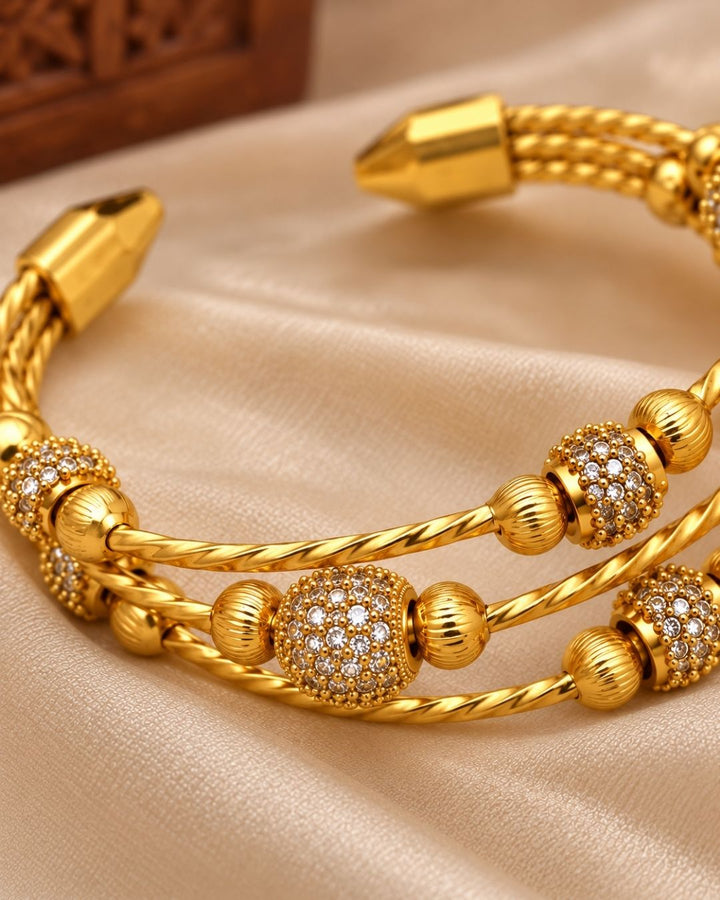 Gold plated adjustable kada for women featuring a triple twisted design, decorative gold beads, and sparkling white stone embellishments.