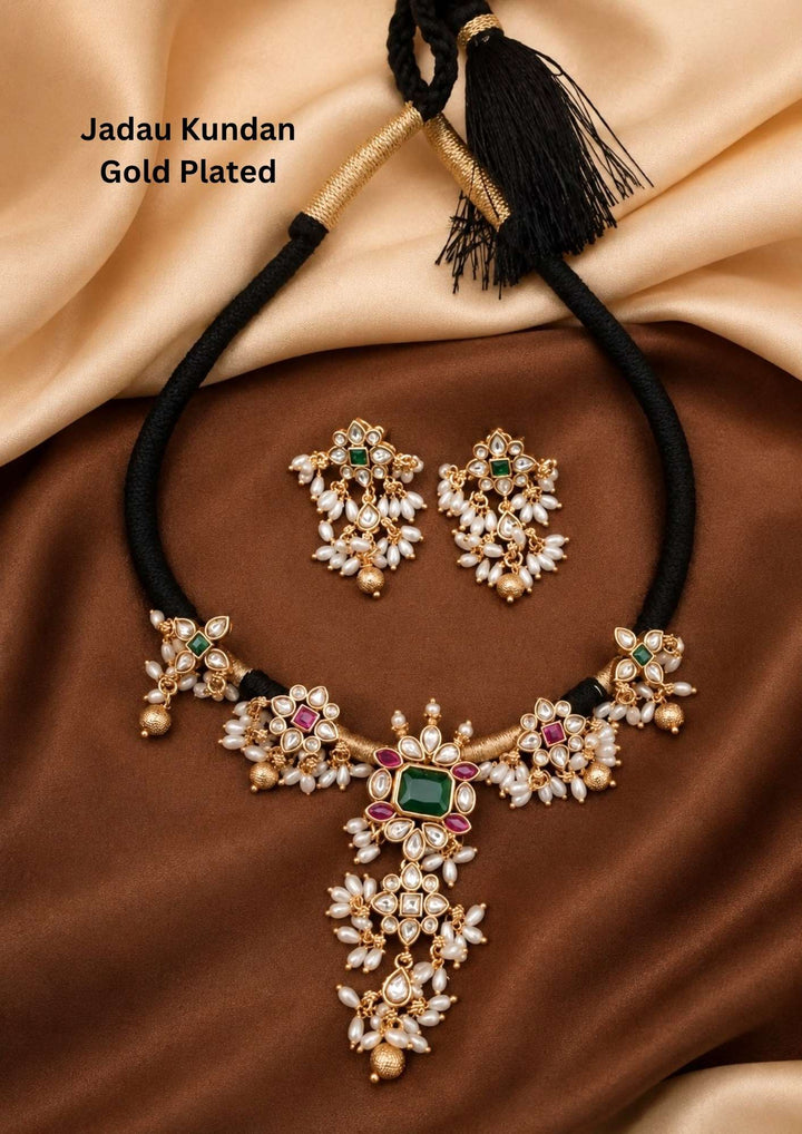 Gold plated short necklace and matching hanging earrings set featuring a black thread cord attached to a structured center piece. The necklace is designed with flower-shaped side links and a square green stone center pendant surrounded by small red and white stones. The lower section of the necklace and earrings is decorated with dense clusters of small hanging white pearls, providing a simple, classic look suitable for festivals and family functions.