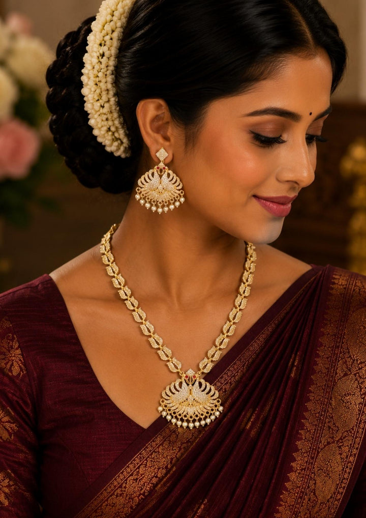South Indian Woman wearing Premium gold plated jewelry set featuring a medium-length link chain decorated with fine white zircon stones and a grand twin peacock pendant arranged in a fan-like wing design. The centerpiece is highlighted with tiny ruby red stone eyes and a delicate fringe of hanging white pearls at the base. It comes complete with matching large peacock drop earrings, crafted with skin-friendly quality