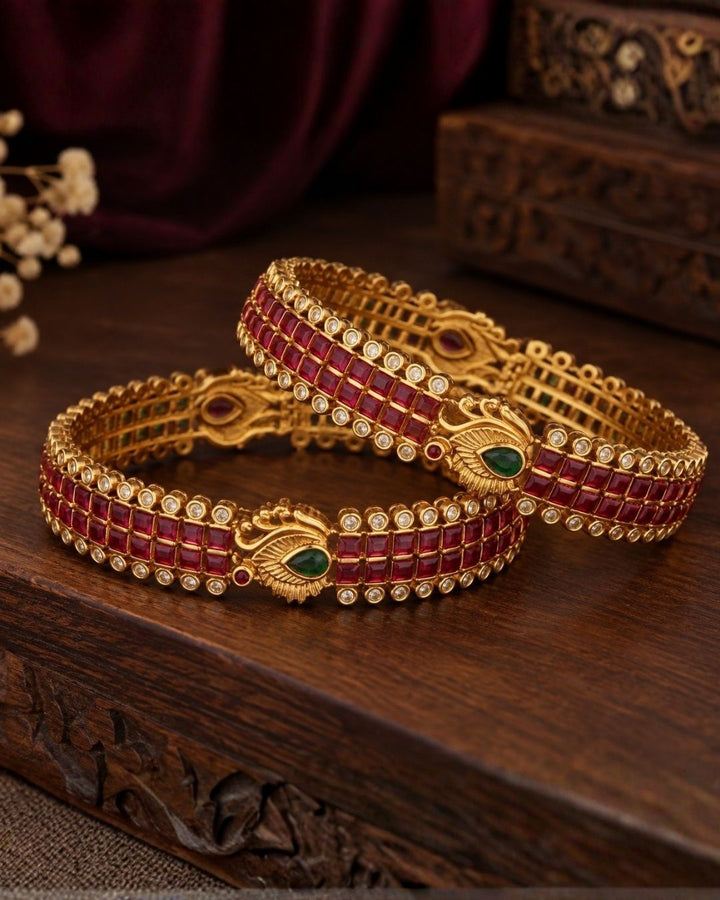 Premium gold-plated real Kemp peacock bangles designed with multiple rows of square-cut red Kemp stones, with white crystal borders, and an ornate golden peacock design centered with a water drop green stone. It is lightweight, skin-friendly, and anti-fade bangles are perfect for poojas, festive wear, and dailywear.