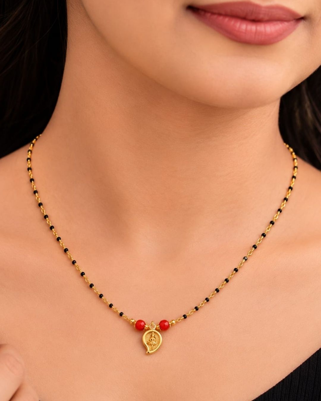 A gold-plated Mangalsutra necklace with red beads and a small gold pendant, displayed on a dark burgundy jewelry bust. Text in the upper left reads 'Mangalsutra 18k Gold Plated'.
