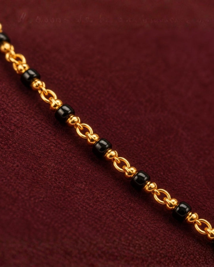 A gold-plated Mangalsutra necklace with red beads and a small gold pendant, displayed on a dark burgundy jewelry bust. Text in the upper left reads &