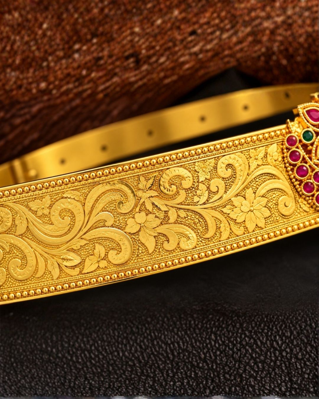 Beautiful gold plated hip belt featuring an intricately engraved wide band with a floral gemstone centerpiece accented with ruby, green, and white stones. Perfect for traditional wear, weddings, festive occasions, bridal styling, parties, and gifting.