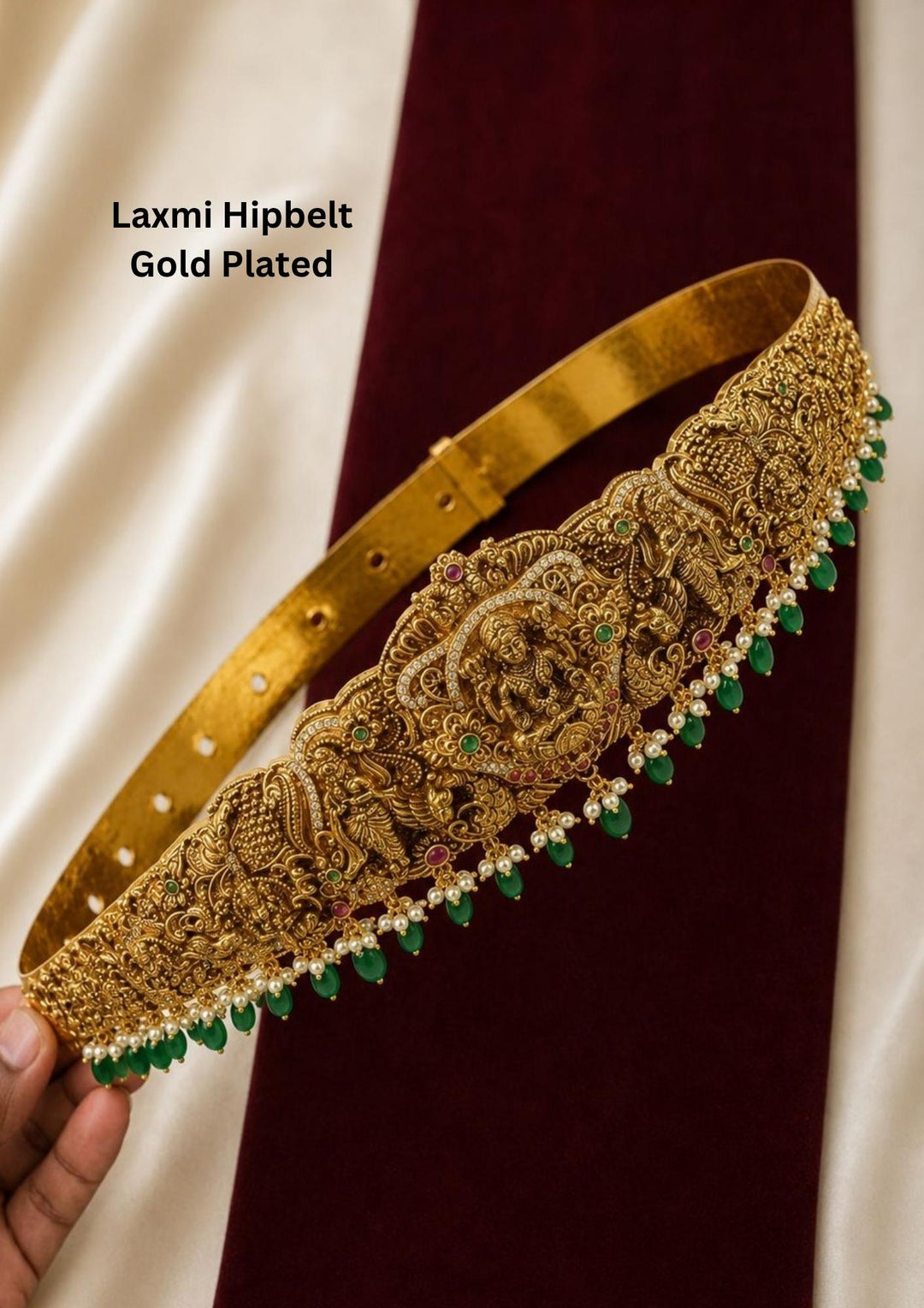 Antique gold plated temple hipbelt or vaddanam, featuring an adjustable solid band that transitions into a highly detailed, broad front panel. The central carving beautifully depicts Goddess Lakshmi seated on a lotus, flanked by dancing peacocks and structural floral archways. The lower border is decorated with a delicate fringe of white pearls and emerald green beads.
