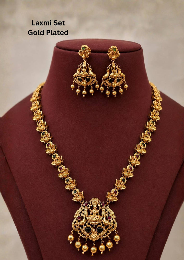 An exquisite antique gold-plated long temple haaram set featuring a prominent Goddess Laxmi pendant. The large, detailed pendant shows the deity seated on a lotus flanked by peacocks, encrusted with ruby-red and emerald-green stones, and finished with hanging textured gold bead balls. Perfect for for bridal and festive wear. It is anti tarnish and skin friendly