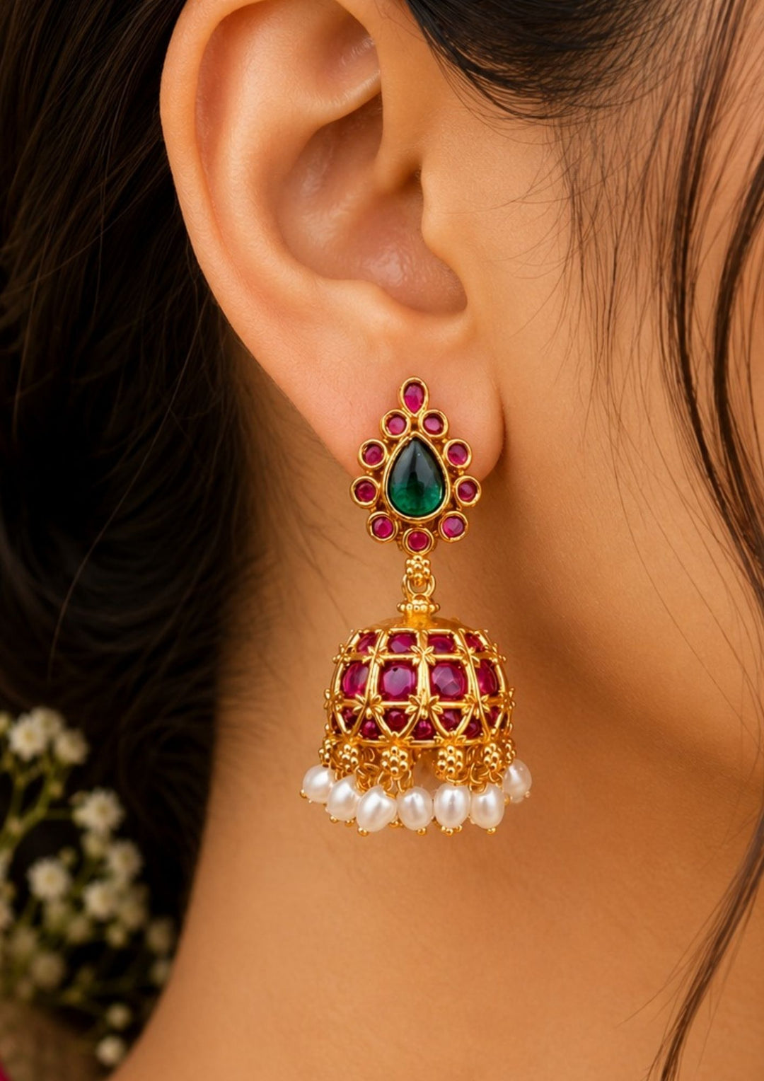Close-up of South Indian  Women ear styled with Jadau Kundan gold-plated earrings. The earrings feature exquisite craftsmanship with embossed traditional deities, openwork filigree, and a vibrant combination of pink, green, and white stones, ideal for bridal and festive jewelry collections.